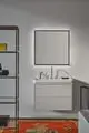 EinWaschtisch mit integrierter Ideal Standard Armatur, zwei Kreuzgriffen und modernem Design. Über dem Waschtisch befindet sich ein beleuchteter Spiegel. Robuste Regalstruktur im Hintergrund.